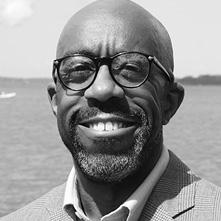 Headshot of a Black man with glasses smiling and standing in front of a body of water